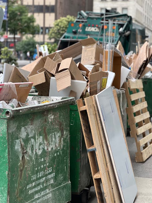 A large green waste disposal skip filled with assorted cardboard boxes of various sizes, some flattened and others still partially assembled, alongside wooden pallets leaning against the side. The cardboard exhibits different textures and light brown tones, with some boxes showing printed labels or tape remnants. The skip is situated on a city street, with a background featuring a green refuse collection truck, multi-storey buildings, and a few trees, indicating an urban environment. The scene is illuminated by natural daylight, highlighting the cluttered arrangement of waste materials, which likely result from on-site or private rubbish clearance services. The detail emphasizes the presence of detached packaging items and the general context of rubbish removal or alternative waste handling, with the emphasis on the practical aspects of waste collection in a city setting, as handled by a service like Rubbish Clearance Lambeth.