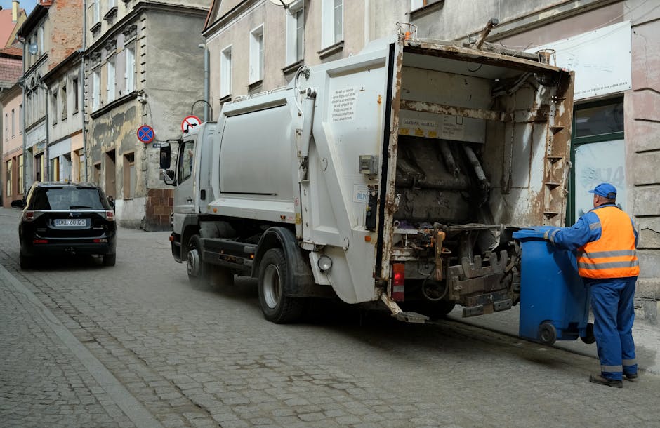 A large green waste disposal skip filled with assorted cardboard boxes of various sizes, some flattened and others still partially assembled, alongside wooden pallets leaning against the side. The cardboard exhibits different textures and light brown tones, with some boxes showing printed labels or tape remnants. The skip is situated on a city street, with a background featuring a green refuse collection truck, multi-storey buildings, and a few trees, indicating an urban environment. The scene is illuminated by natural daylight, highlighting the cluttered arrangement of waste materials, which likely result from on-site or private rubbish clearance services. The detail emphasizes the presence of detached packaging items and the general context of rubbish removal or alternative waste handling, with the emphasis on the practical aspects of waste collection in a city setting, as handled by a service like Rubbish Clearance Lambeth.