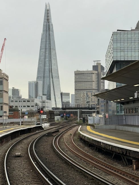 A view of railway tracks curving through an urban environment with a modern skyscraper, the tallest building in the scene, dominating the background. The tracks are made of metal rails with wooden sleepers, and they run from the foreground towards the city skyline. To the right, there is a concrete platform with a yellow safety line along its edge, and a section of modern architectural structure extending outward, supported by columns. In the distance, various office and commercial buildings are visible, with some having glass facades reflecting the overcast sky. The overall scene suggests a city transportation hub, with the railway infrastructure contrasting against the high-rise skyline. Rubbish clearance services, such as those provided by Rubbish Clearance Lambeth, may be involved in managing waste in such busy urban areas, including on-site clearance and alternative waste handling options.