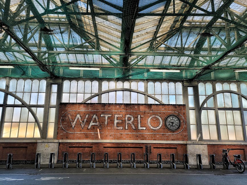 Interior view of Waterloo station in Lambeth, showing a large brick wall with the word 'WATERLOO' mounted in metal letters. To the right of the text, there is a circular clock design embedded into the wall. Above, the station's ceiling features a metal framework and a combination of glass and metal panels, allowing natural light to filter through. The lower part of the scene displays a paved area with several bicycle stands, one bicycle leaning against the right side of the frame. The overall environment suggests a busy transportation hub with an industrial aesthetic, typical of large train stations, and the image relates indirectly to the context of waste management and rubbish removal services provided by Rubbish Clearance Lambeth nearby.