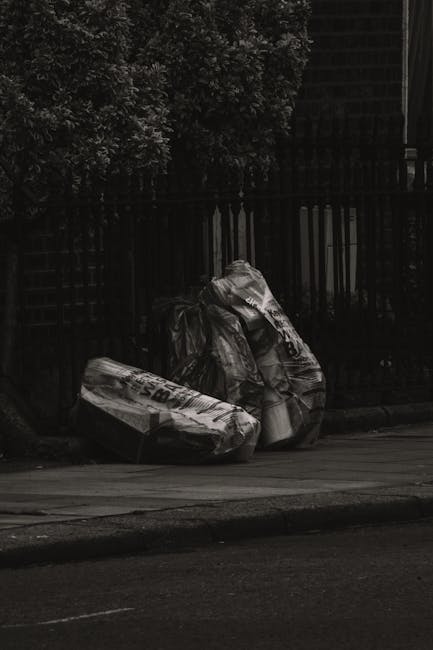 Two large black plastic rubbish bags, filled and tied at the top, are placed on the pavement nearest a black metal fence with vertical bars. One bag appears slightly more crumpled than the other, and both sit on a narrow strip of concrete, with a dark road surface in the foreground. Behind the fence, dense foliage from trees or bushes is partially visible, creating a contrasting natural background against the urban setting. The scene is captured in subdued lighting, potentially early morning or late evening, with no visible presence of people or vehicles. The arrangement suggests the bags are awaiting collection, fitting into the context of independent waste handling services such as those provided by Rubbish Clearance Lambeth, which handles various types of household or commercial rubbish removal within Lambeth. This outdoor setting exemplifies typical rubbish disposal practice outside residential or commercial premises, emphasizing the need for professional waste clearance solutions to manage refuse efficiently and responsibly.