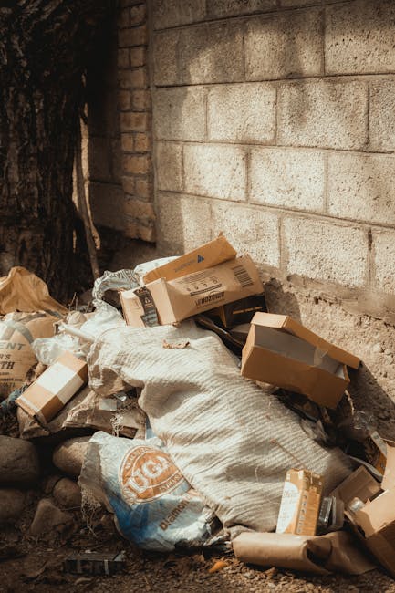 A pile of discarded cardboard boxes, some flattened and others partially assembled, is gathered against a rough concrete brick wall in an outdoor setting. The boxes vary in size and are brown, with some showing printed labels or barcodes. Among the cardboard, there are crumpled paper and plastic bags, including a branded plastic bag with orange and white graphics. A white fabric sack or large cloth bag is also part of the debris, partially covering some of the boxes and materials. The ground beneath the waste consists of earthy soil and small rocks, with a dark tree trunk visible on the left side of the image, indicating an exterior environment. The scene is illuminated by natural daylight, casting soft shadows on the waste and wall, presenting a typical example of unsorted rubbish that a professional waste collection service such as Rubbish Clearance Lambeth might handle as part of a private rubbish clearance or on-site waste removal operation. The setting appears to be an alleyway or side yard, with the wall providing a clear background for the accumulated refuse, illustrating typical residential or small-scale waste accumulation for disposal or recycling.