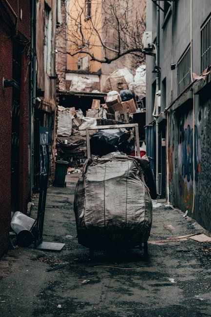 A narrow urban alleyway filled with a large pile of mixed waste and discarded materials positioned against a backdrop of cluttered cardboard boxes, plastic bags, and debris stacked up on a raised surface at the end of the alley. In the foreground, a substantial black trash bag or sack is strapped onto a wheeled trolley or hand truck, suggesting an ongoing rubbish clearance process by Rubbish Clearance Lambeth. The alley is flanked by multi-storey buildings with metal security bars on windows and graffiti on walls, and the surface of the ground appears worn and stained, with patches of dirt and scattered trash. Bare tree branches are visible overhead, indicating an outdoor setting, and the overall atmosphere is dimly lit, reflecting typical conditions of a behind-building rubbish accumulation site. This scene illustrates a typical refuse collection environment where private waste disposal or on-site clearance is undertaken to manage accumulated rubbish in an urban setting.
