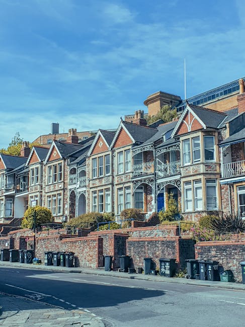 A row of Victorian-style terraced houses with detailed decorative ironwork on each small front balcony and white wooden window frames. The buildings feature red brick walls combined with lighter stone accents and triangular gabled roofs, some with small dormer windows. The houses are elevated above the pavement, accessible via short flights of stairs bordered by low brick walls. In front of the houses, there is a narrow sidewalk lined with neatly trimmed bushes and small trees. Below the brick wall, several black and green wheelie bins are positioned along the curb, indicating waste collection readiness. The street surface is paved with asphalt and has minimal traffic markings. The scene is captured on a clear day with a bright blue sky dotted with light clouds, suggesting a peaceful residential area. The visible environment and waste disposal elements subtly relate to independent rubbish removal and alternative waste handling options, aligning with the context of rubbish clearance services.