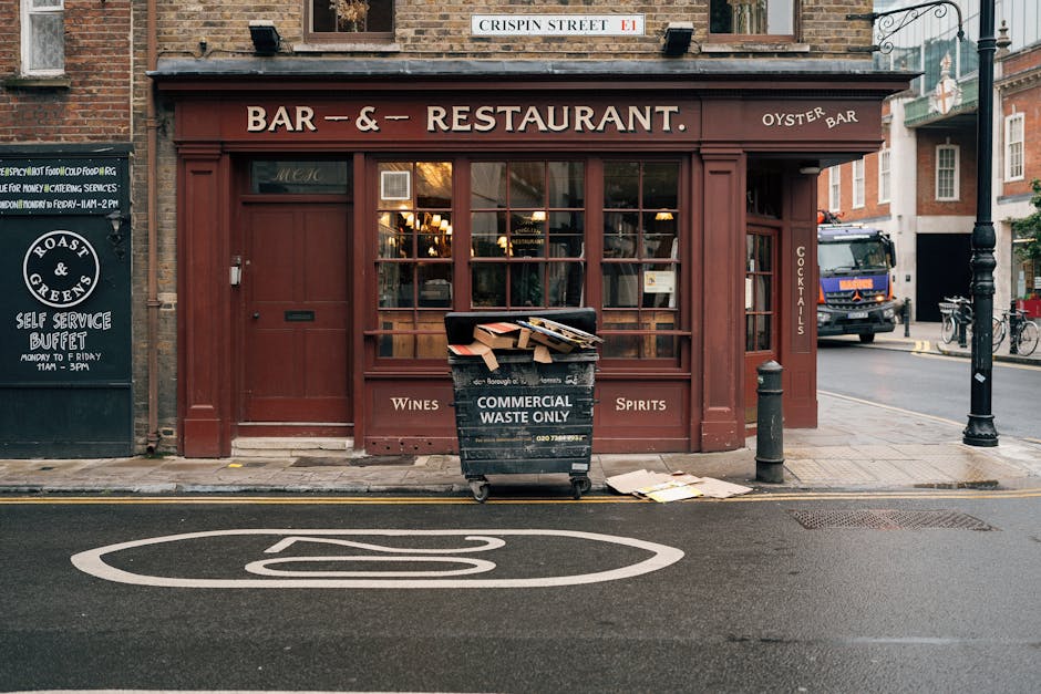 In front of a red-brick restaurant with a large window and wooden paneling, there is a black commercial waste bin labeled 'Commercial Waste Only' positioned on the sidewalk. The bin contains flattened cardboard boxes and some scattered paper or cardboard pieces lying on the ground nearby. The restaurant signage indicates it is a bar and restaurant serving wine, spirits, and cocktails, with an oyster bar mentions. To the left, a blackboard sign provides information about a self-service buffet, and to the right, a black bollard is visible near the curb. A street sign labeled 'Crispin Street' is mounted above the building. The scene is outdoors with typical urban street elements including a parked truck in the background, bicycles leaning against a railing, and street lamps, all under natural daylight, providing context for private waste handling and rubbish removal services in an active commercial area. The setting emphasizes the typical placement of waste collection on public pavements as part of routine rubbish clearance.