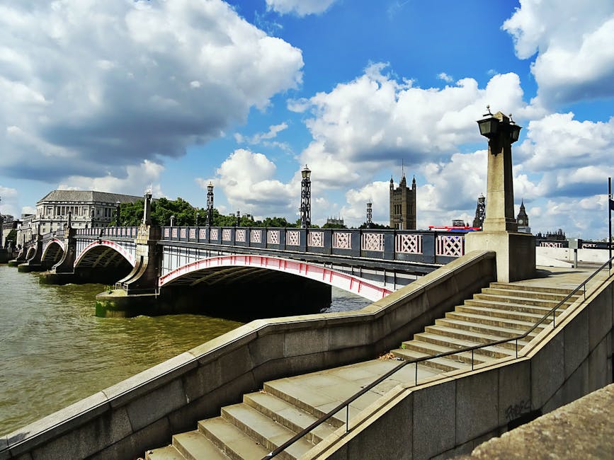 The image depicts a section of the Westminster Bridge spanning the River Thames in London, with the steps of a riverbank promenade in the foreground. The bridge features decorative ironwork with a white and pink color scheme, supported by stone arches extending into the water. On the right side, a large stone pillar topped with a classic lamppost marks the staircase leading down to the riverbank. In the background, the Houses of Parliament and Westminster Abbey are visible, with their historic Gothic Revival architecture rising against a partly cloudy sky filled with large, fluffy clouds and patches of blue. The scene includes the river's muddy-green water and reflections of the bridge and nearby structures, creating a picturesque yet neutral setting typical of central London. This view aligns with images often associated with London landmarks and urban scenery, complementing content about city travel or outdoor sightseeing, and can support services dealing with waste or rubbish collection near iconic sites through natural environmental context.