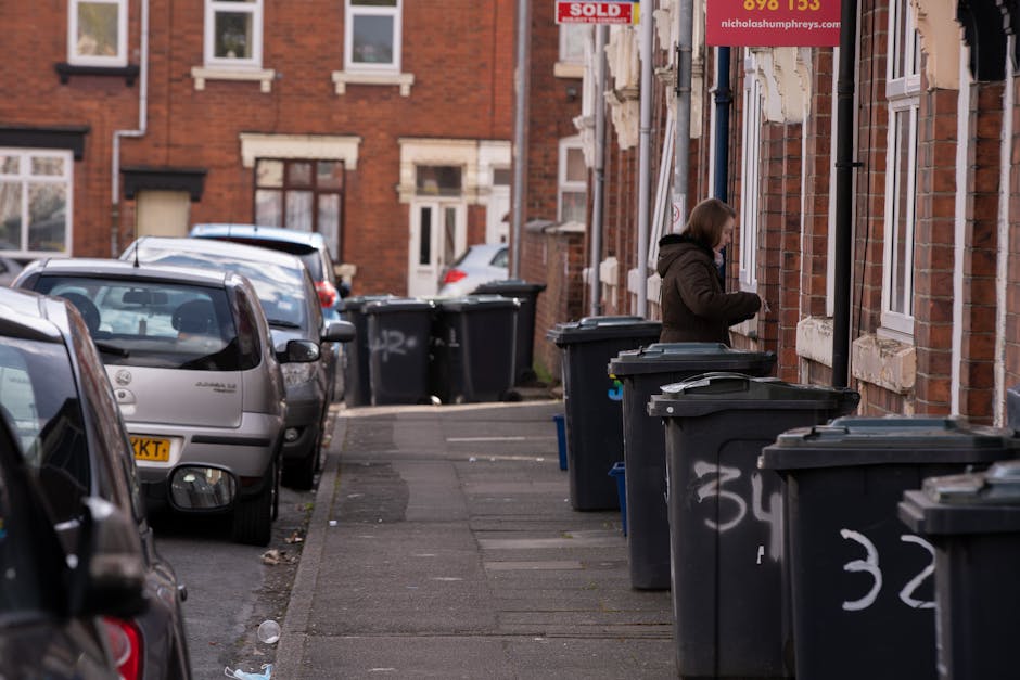 A woman standing on an urban sidewalk next to a row of large dark grey wheelie bins with white numbering, situated against the brick facade of residential buildings. The bins are made of plastic with hinged lids, some slightly open, revealing piled rubbish inside. The sidewalk is narrow, with a curb separating it from parked cars that occupy the street, including a silver hatchback close to the viewer. Painted graffiti is visible on the lower parts of the bins, with a background of red-brick townhouses featuring white-framed windows and some signage, including a red advertisement for a property sale. Natural daylight illuminates the scene, casting minimal shadows. The environment appears typical for a residential area, with the bins arranged for collection or private waste disposal, reflecting an aspect of independent rubbish collection services. Rubbish clearance company Rubbish Clearance Lambeth occasionally handles such collections for local residents or businesses. The overall composition emphasizes the arrangement and materials of waste containers against the urban setting, suitable for an article about rubbish collection or waste management in Clapham High Street areas.