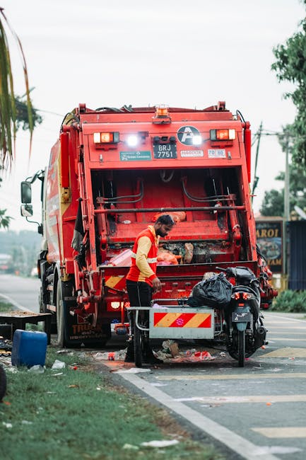 A sanitation worker wearing a red and yellow uniform is operating a red garbage truck on a street, with the rear hatch open, revealing mechanical components and collection mechanisms. The worker isusing a tool to collect waste from a black plastic bag placed on a metal platform attached to the back of the vehicle. The truck is parked partially on the pavement, with debris and waste materials visible on the ground near the collection area. In the background, there are trees, power lines, and a glimpse of storefronts or signage, indicating an urban or suburban environment. The scene appears to be during daylight with overcast skies, providing soft, even lighting. The image illustrates the process of rubbish collection, relevant to private waste management services such as those offered by Rubbish Clearance Lambeth, highlighting on-site waste handling and collection methods typical of independent rubbish removal services.