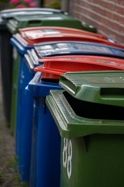 A row of plastic rubbish bins lined up outdoors along a pavement beside a brick wall, with the bins in various colours including green, red, blue, and dark green. The bins are fitted with lids, some of which are slightly open, revealing the interior space for waste. The green bin in the foreground shows a white recycling symbol on its side, indicating its designated waste type. The surface of the bins appears to be made of durable, textured plastic, with a slightly glossy finish, reflecting some natural light. The bins are positioned closely together, creating a slight curve in the line extending towards the background, where more bins are visible, gradually fading out of focus. The setting suggests an urban or residential area and exemplifies common private waste collection methods, potentially serviced by local rubbish clearance providers such as Rubbish Clearance Lambeth, who handle alternate disposal and on-site rubbish removal services in the Lambeth area.