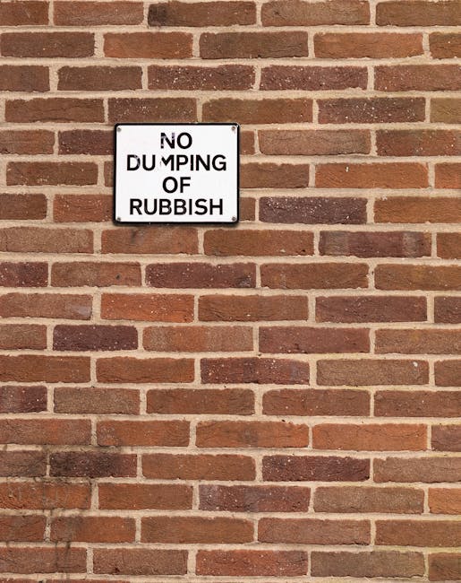 A rectangular white sign with black text that reads 'NO DUMPING OF RUBBISH' is mounted on a red brick wall. The brick wall features traditional Flemish bond pattern with reddish-brown bricks and light grey mortar joints, some bricks showing slight weathering. The sign is positioned near the top left quadrant of the image, with the bricks extending horizontally and vertically across the entire background. The wall appears to be part of an exterior structure, possibly a boundary wall or building façade, with no other objects or environmental features visible. The lighting is even, indicating daylight, and the overall scene conveys a notice aimed at preventing unauthorised waste disposal in this area, supporting the context of private rubbish management or site-specific rubbish removal regulation, as might be relevant for services offered by Rubbish Clearance Lambeth.