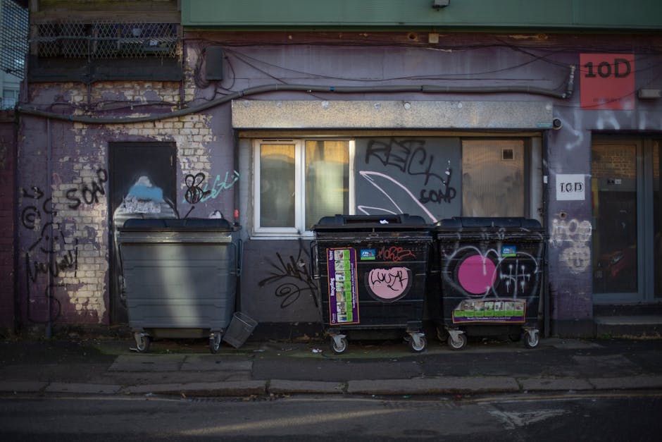 A row of plastic rubbish bins lined up outdoors along a pavement beside a brick wall, with the bins in various colours including green, red, blue, and dark green. The bins are fitted with lids, some of which are slightly open, revealing the interior space for waste. The green bin in the foreground shows a white recycling symbol on its side, indicating its designated waste type. The surface of the bins appears to be made of durable, textured plastic, with a slightly glossy finish, reflecting some natural light. The bins are positioned closely together, creating a slight curve in the line extending towards the background, where more bins are visible, gradually fading out of focus. The setting suggests an urban or residential area and exemplifies common private waste collection methods, potentially serviced by local rubbish clearance providers such as Rubbish Clearance Lambeth, who handle alternate disposal and on-site rubbish removal services in the Lambeth area.