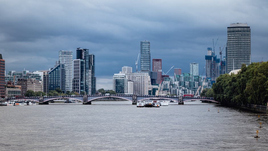 A wide-angle view of a city skyline across a river, featuring numerous modern high-rise buildings with glass facades and varying architectural styles, some under construction with cranes visible. The river in the foreground is calm with a few small boats moored near the banks. The cloudy sky overhead casts a diffused light over the scene. On the right side, a dense row of green trees lines the riverbank, contrasting with the urban environment. In the middle distance, an arched bridge spans the river, connecting the two sides of the city. The overall scene depicts a bustling metropolitan area with a mix of residential, commercial, and office buildings, illustrating the type of urban environment where private waste management or rubbish removal services, such as those offered by Rubbish Clearance Lambeth, can support effective on-site waste clearance for construction, renovation, or general rubbish collection.