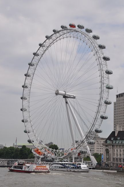 A large observation wheel with a metal framework and numerous enclosed passenger cabins evenly spaced around its circumference, situated on a riverbank or waterfront area. The wheel is supported by a sturdy, angled white structure with a central axle connecting to the main hub. The cabins are mostly glass-fronted, with some showing dark or transparent windows, and are painted in neutral tones, blending with the metallic frame. In the foreground, a white and red river cruise boat with the logo 'citycruises' on its side is sailing on the water beneath the wheel. The background features a cloudy sky and some buildings, including a tall, rectangular structure and a historic-looking building with architectural detailing, indicating an urban setting. The scene captures a typical city riverside environment, where the Ferris wheel serves as a prominent sightseeing attraction, complemented by the nearby water transportation, aligning with services for alternative or independent sightseeing and recreational boat trips in the area.