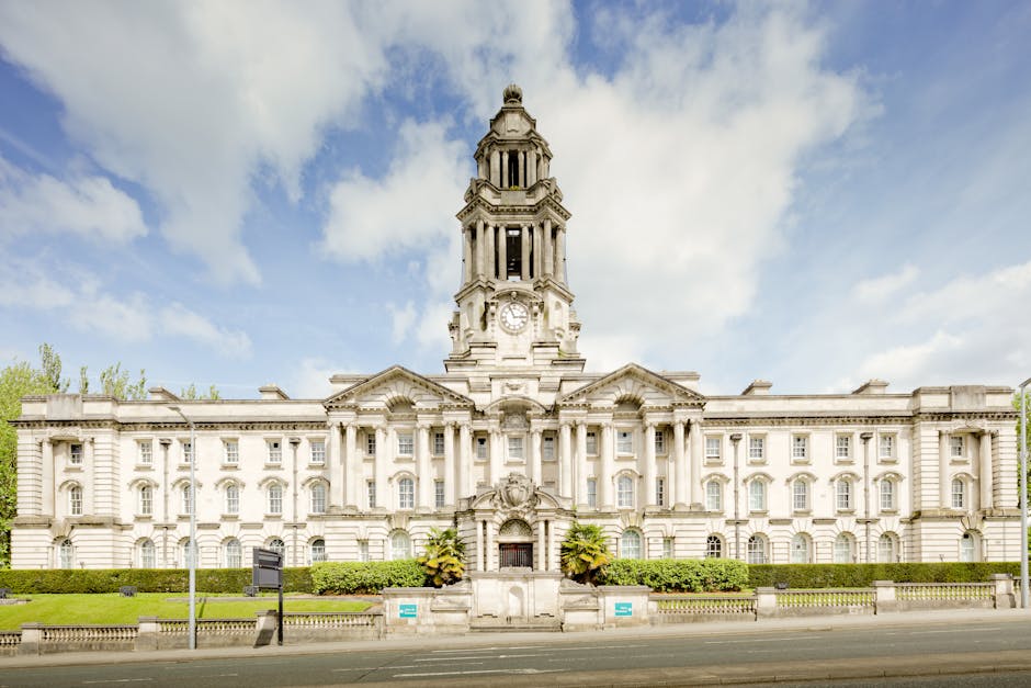 A large, historic government building made of light-colored stone, featuring detailed classical architectural elements such as columns, arches, and decorative sculptures. The central tower rises above the structure, topped with a domed or sculpted element, and contains a clock face visible near the top. The façade is symmetrical with multiple windows, some arched and others rectangular, framed by ornate stonework. In front of the building, a low stone wall and neatly trimmed green hedges run along the pavement, with a few small trees and potted plants situated near the entrance. The scene is set outdoors during daylight, with a partly cloudy blue sky overhead and natural light illuminating the stone details. The setting appears urban, with a street running directly in front of the building, suggesting a city or town centre location, and subtly reflects context related to municipal or government services, complementing the theme of property and waste management services involved in public or private waste handling or site clearance activities managed by companies like Rubbish Clearance Lambeth.